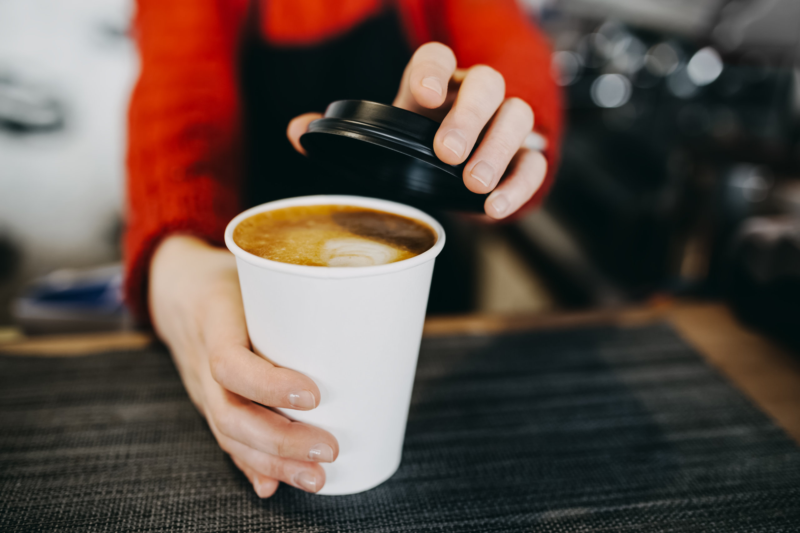 Barista in apron is holding in hands hot cappuccino in white takeaway paper cup. Coffee take away at cafe shop Barista in apron is holding in hands hot cappuccino in white takeaway paper cup. Coffee take away at cafe shop